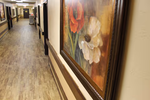 A long, well-lit hallway in a nursing and rehabilitation facility with wood-patterned flooring, beige walls, handrails, and framed floral artwork hanging on the wall.