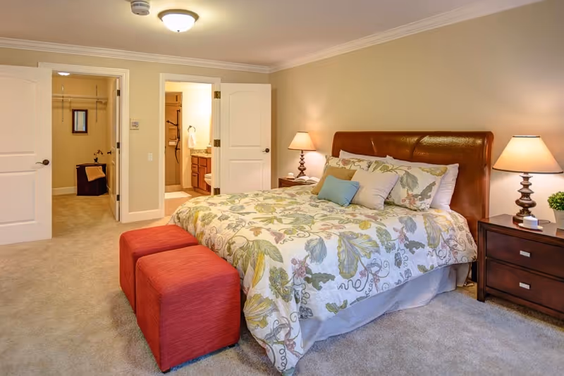 A cozy bedroom with a large bed featuring a floral patterned comforter and multiple pillows. Two red ottomans are placed at the foot of the bed. There are two bedside tables with lamps on each side of the bed. The room has beige walls and carpeted floor. Two open doors reveal a walk-in closet and a bathroom with a vanity and mirror.