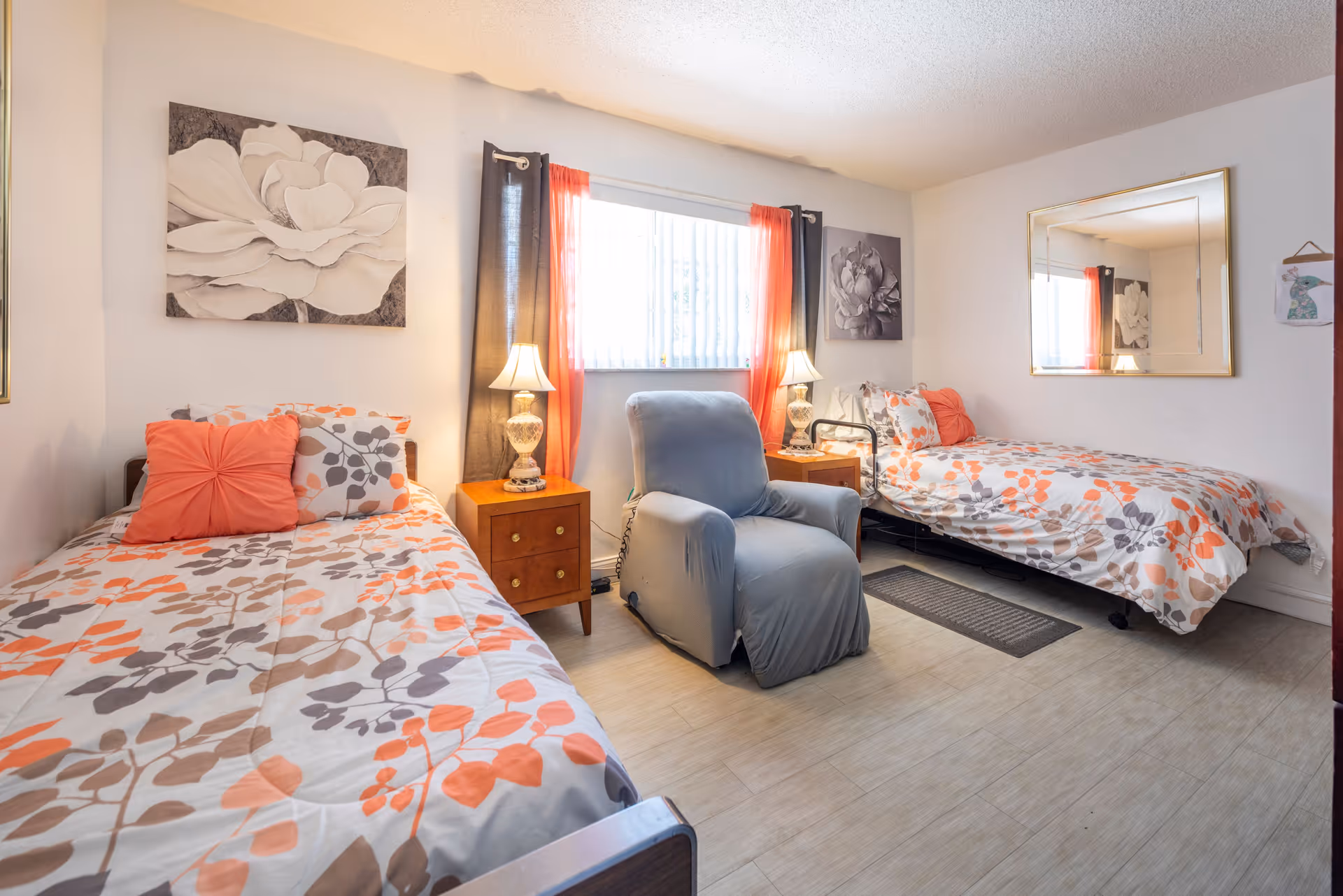 A bright bedroom with two single beds featuring floral-patterned bedding in shades of orange, gray, and white. Between the beds are two wooden nightstands with decorative lamps. A comfortable gray armchair is positioned in front of a window with vertical blinds and orange and dark gray curtains. The walls are decorated with large floral artwork and a large rectangular mirror.
