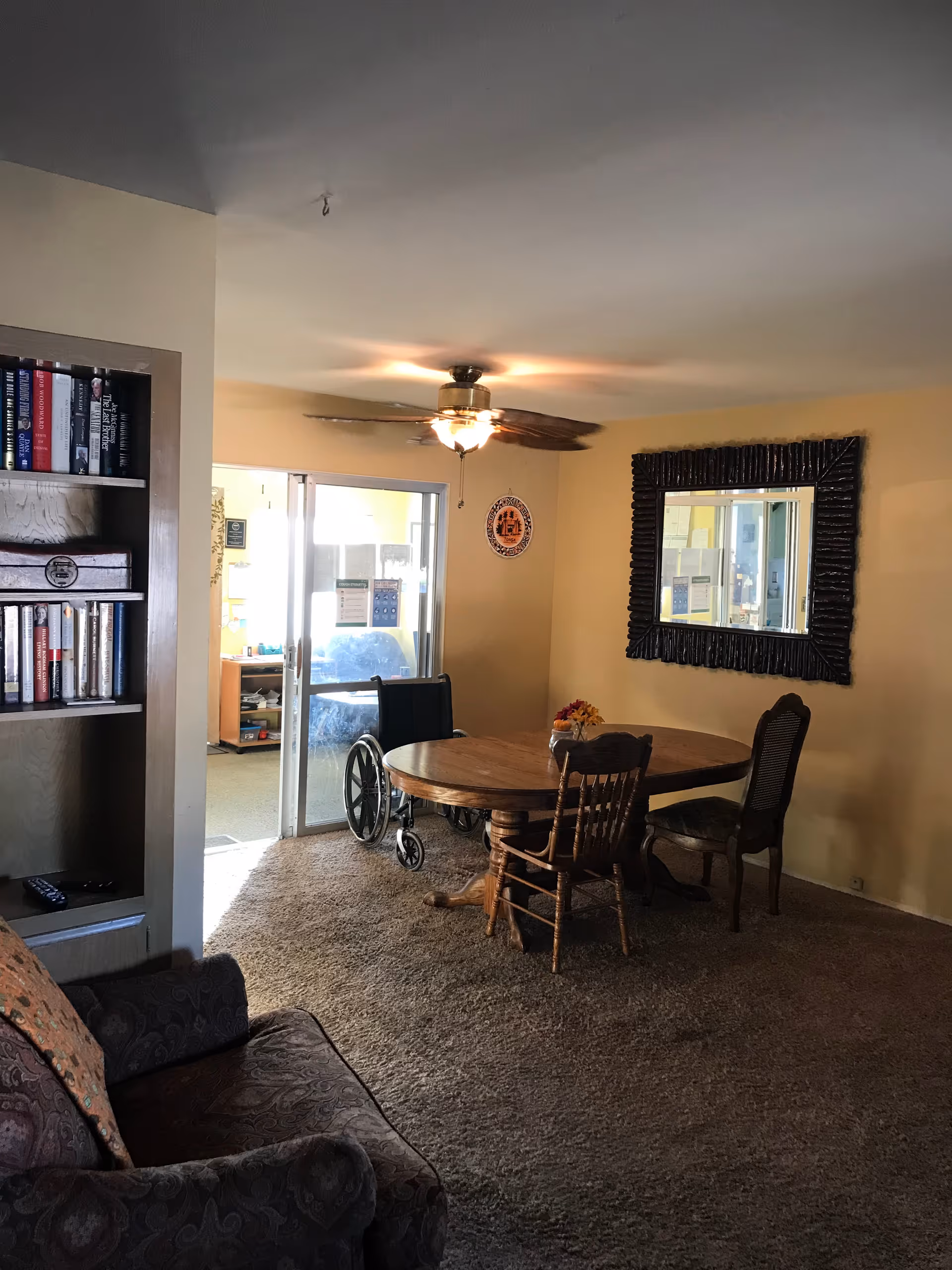 Small dining area with a round wooden table and chairs, a wheelchair, bookshelf, ceiling fan and a large wall mirror.