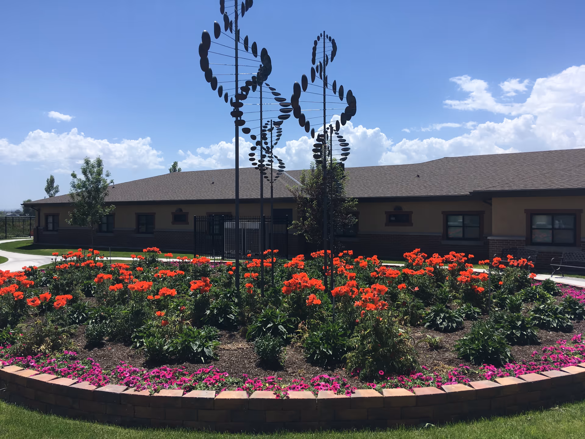 A circular flower bed with vibrant red and purple flowers in front of a single-story building under a blue sky with some clouds. In the center of the flower bed are tall metal sculptures resembling DNA helices. There is a paved walkway surrounding the flower bed and some trees near the building.