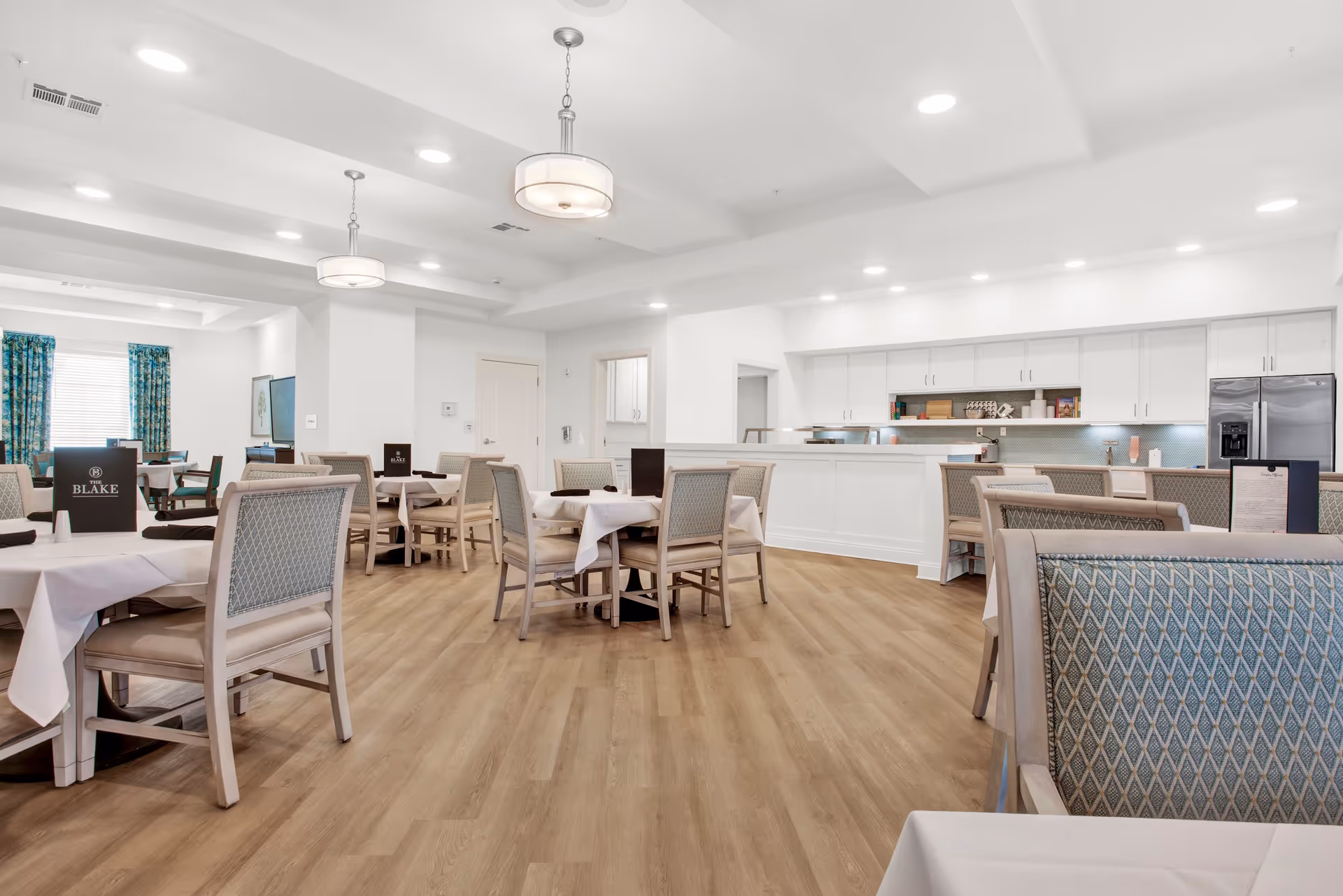A bright and spacious dining room with multiple tables covered in white tablecloths and surrounded by cushioned chairs. The room features light wood flooring, white walls, and ceiling with recessed lighting and hanging light fixtures. In the background, there is a kitchen area with white cabinetry, a stainless steel refrigerator, and a countertop. Blue patterned curtains cover the windows on the left side of the room.