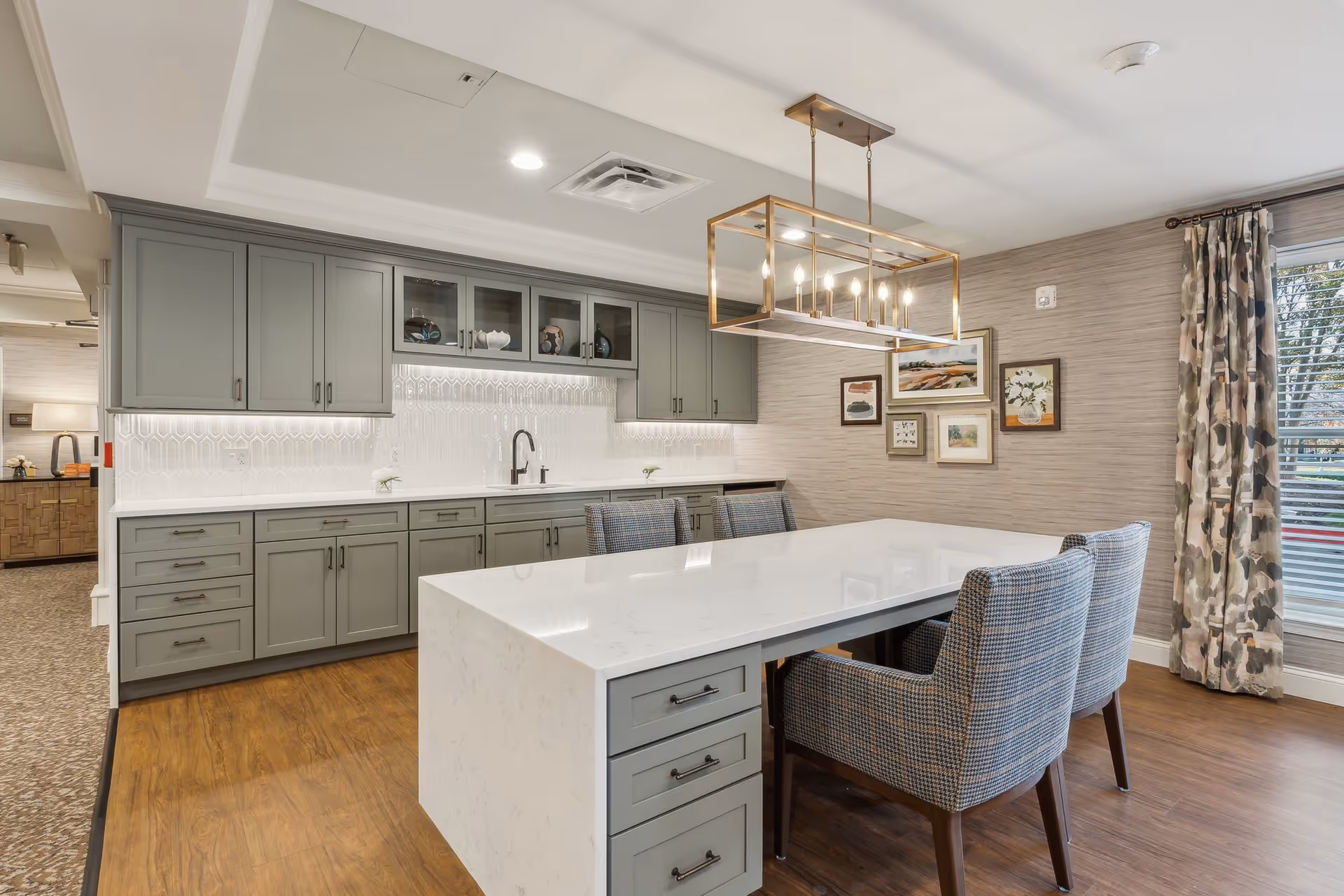 A modern kitchen and dining area with gray cabinets, a white marble countertop island, four upholstered chairs, a geometric pendant light fixture, framed artwork on the wall, and a window with patterned curtains.