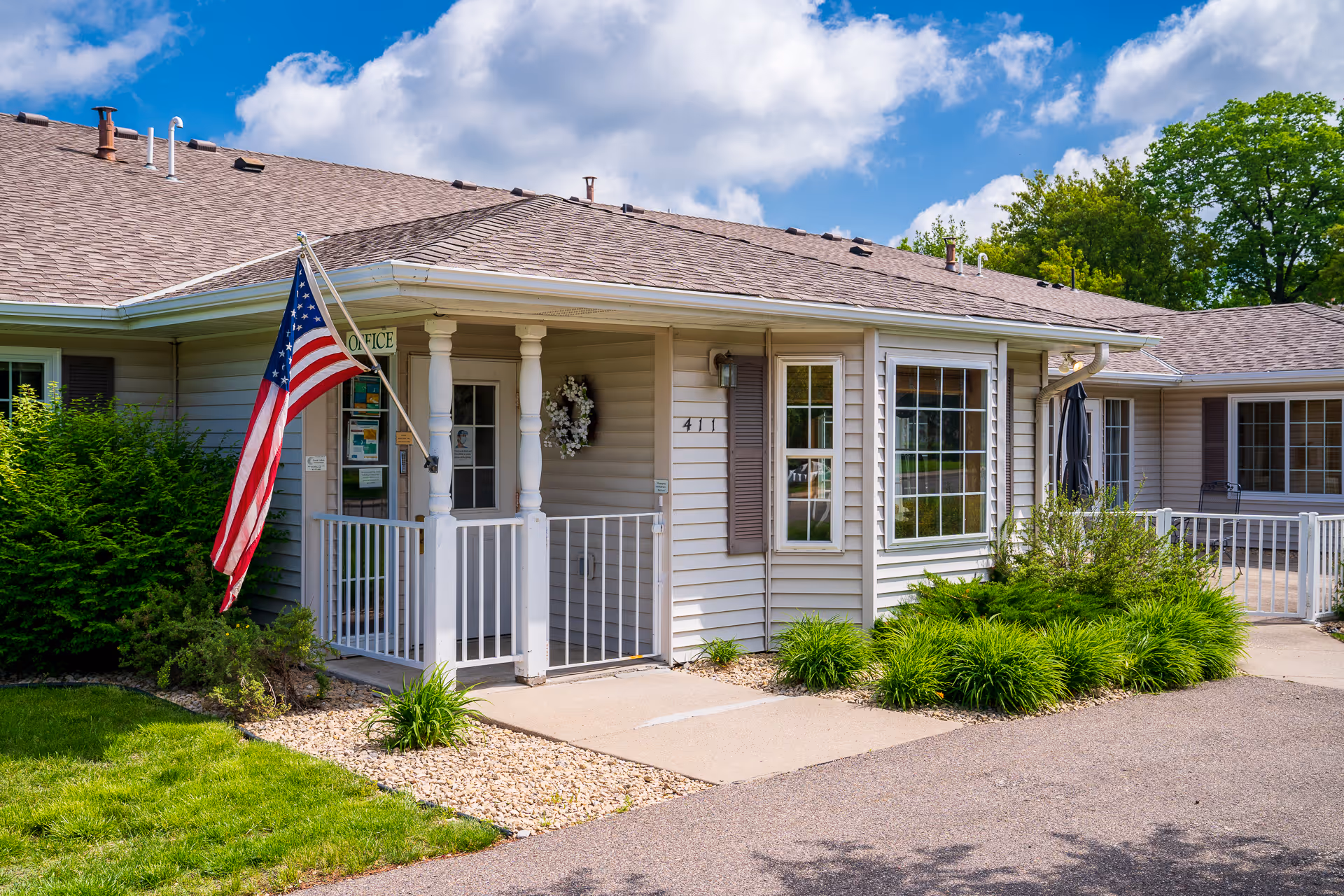 Exterior view of a single-story building with beige siding and a brown shingled roof. The entrance has a small porch with white railings and columns, an American flag mounted on a pole, and a sign that reads 'Office'. There are green bushes and plants around the building, and the sky is partly cloudy with blue patches.