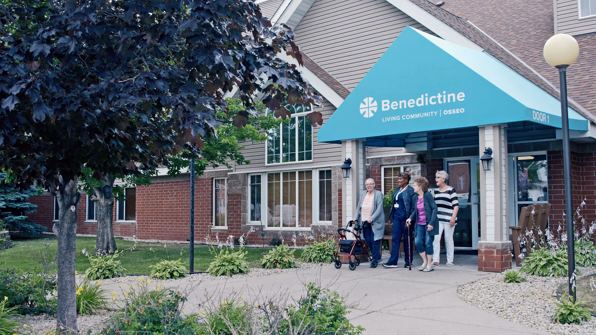 Front entrance of Benedictine Living Community–Osseo with a teal canopy and four women standing on the sidewalk, one with a walker.