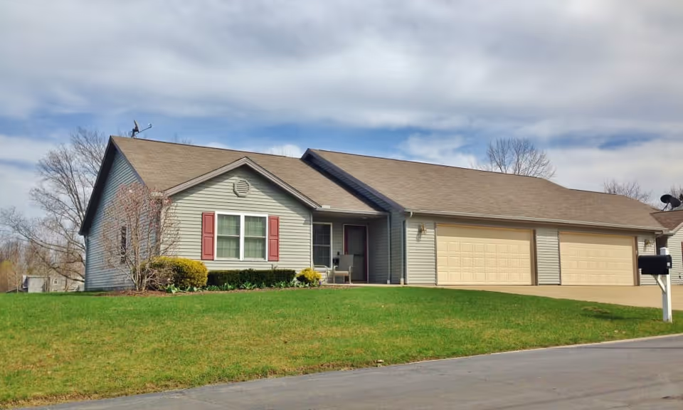Single-story ranch-style house with an attached three-car garage, front lawn, and mailbox under a partly cloudy sky.