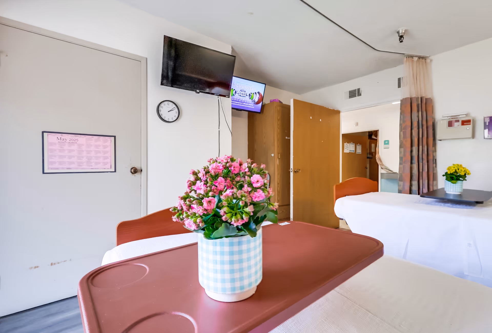 Interior view of a senior living facility room with two beds, a brown overbed table with a potted pink flower, a wall-mounted TV, a clock, an open wooden door, and a curtain partially covering the entrance to another room.