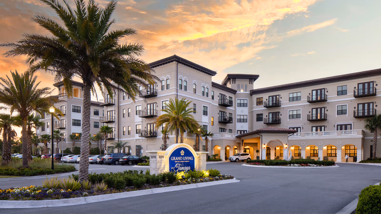 Exterior view of a large, multi-story senior living facility named Grand Living At Tamaya during sunset. The building features balconies, arched windows, and a covered entrance. Palm trees and landscaped greenery surround the driveway and parking area in front of the building.