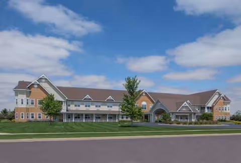 A large senior living facility with light-colored facade and gabled roofs sits on a manicured lawn.