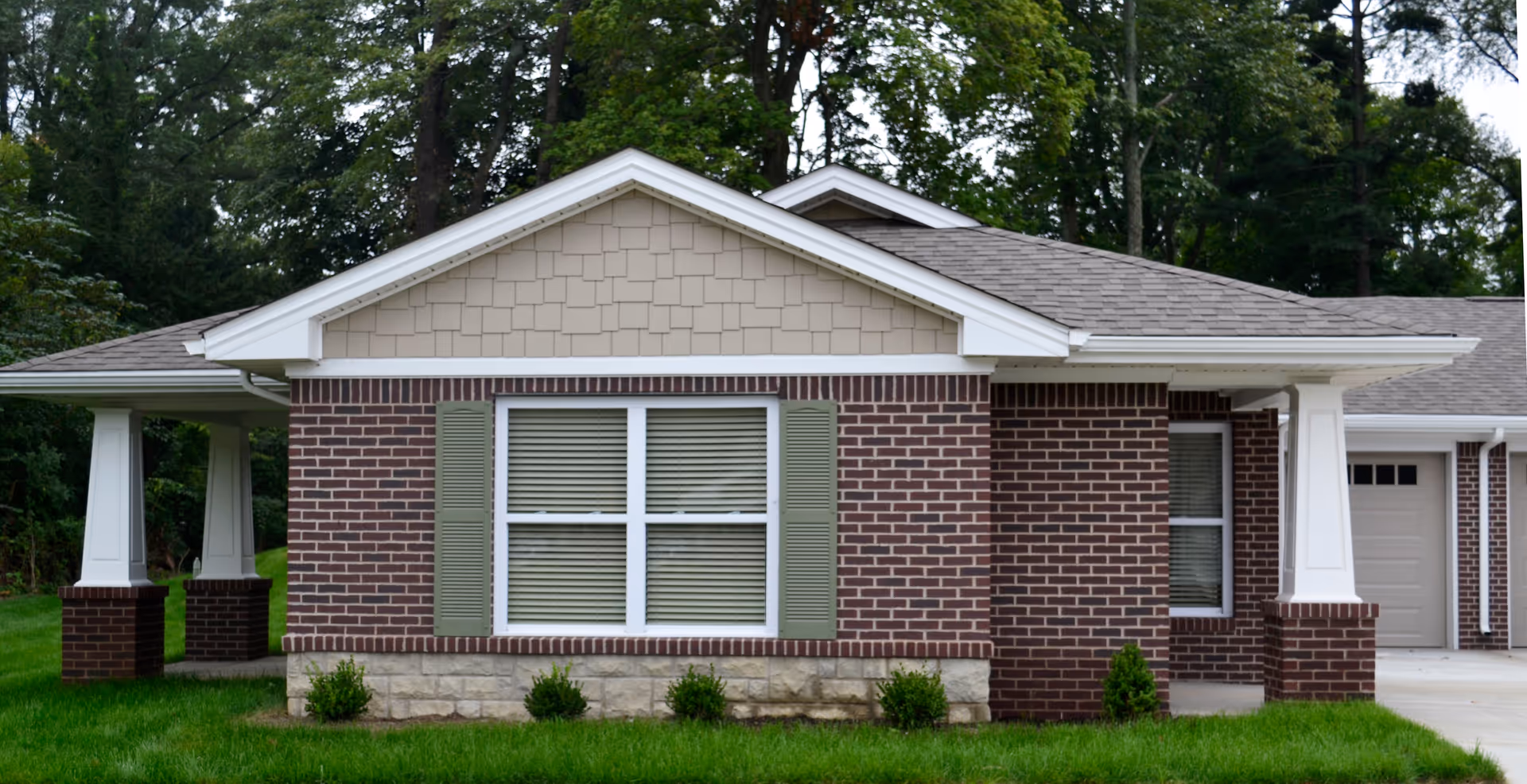 Exterior view of a single-story brick building with a gabled roof, green shutters on the windows, white columns supporting a covered porch, and a well-maintained green lawn in front. Trees are visible in the background.