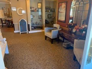Interior view of a senior living facility lobby or common area with carpeted floor, two armchairs, a wooden console table with a flower arrangement, a large mirror, framed artwork on the walls, and a display cabinet with various items inside.