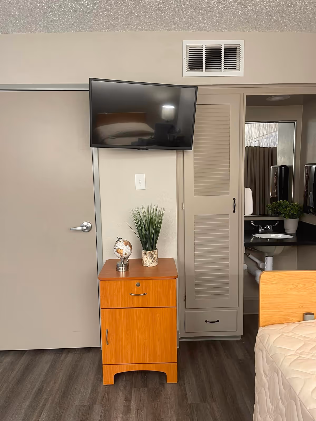A corner of a nursing facility room featuring a mounted flat-screen TV above a wooden cabinet with a small globe and a potted plant on top. To the right, there is a partial view of a bed with a beige quilt and a sink area with a mirror and another potted plant. The room has wood-style flooring and neutral-colored walls.