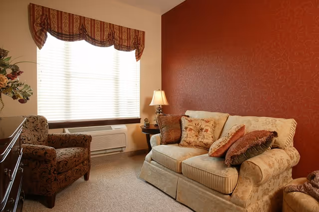 A cozy living room with a beige patterned sofa adorned with multiple decorative pillows, a matching armchair with a dark floral pattern, a wooden side table with a lamp, and a large window with white blinds and a red and gold valance. The wall behind the sofa is painted red with a subtle floral pattern.