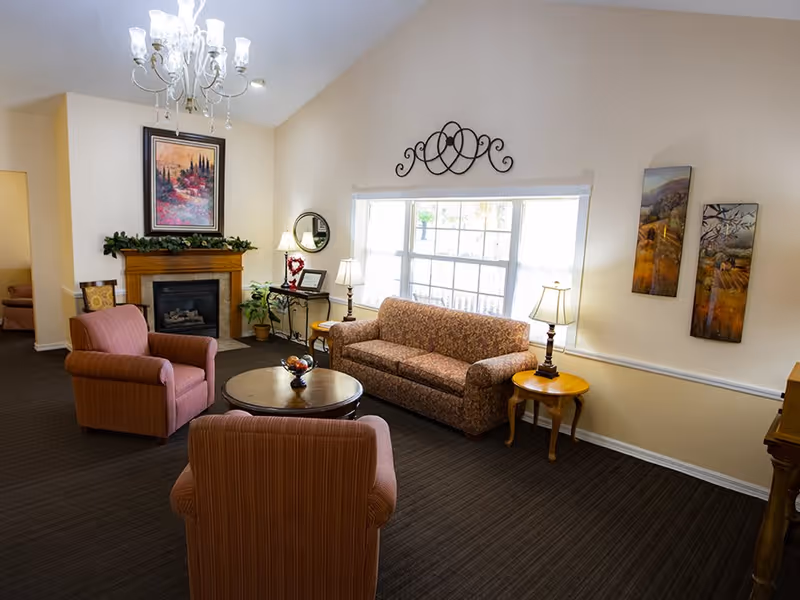 A cozy living room with a patterned sofa and two striped armchairs arranged around a round wooden coffee table. The room features a fireplace with a framed painting above it, a chandelier hanging from the ceiling, and several table lamps providing warm lighting. There are decorative wall hangings and a large window letting in natural light.