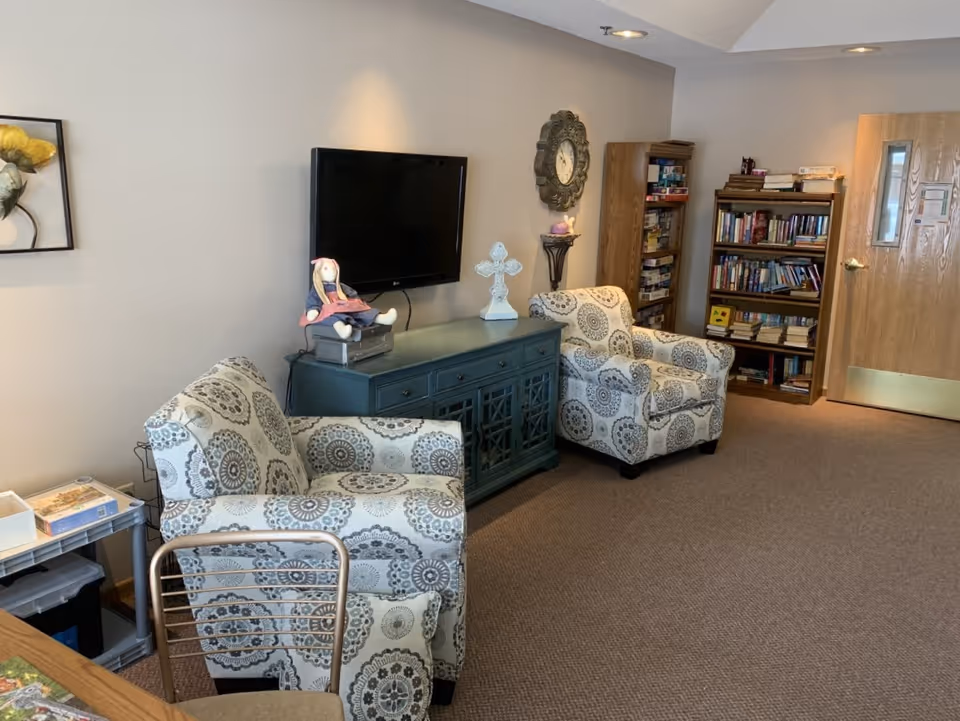 A cozy living room area with two patterned armchairs, a blue cabinet with a doll and a decorative cross on top, a wall-mounted flat screen TV, a decorative wall clock, and two bookshelves filled with books and board games. The room has beige walls and carpeted floor.