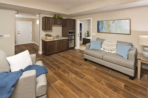 A cozy living room area with a gray sofa adorned with blue and beige pillows, a matching armchair with a blue throw blanket, a wooden side table with a lamp, and a small kitchenette with dark wood cabinets and a built-in microwave and oven. The floor is covered with wood-look tiles, and there is a colorful abstract painting on the wall.