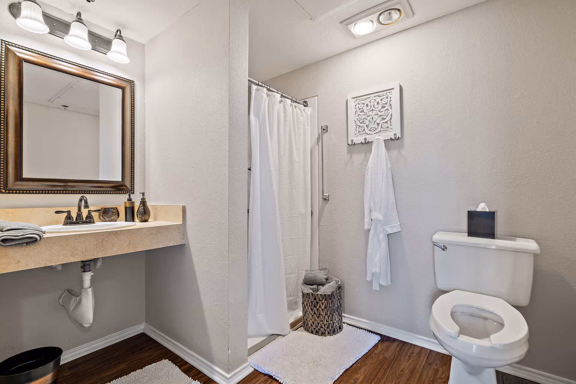 A clean bathroom featuring a white toilet with an open seat, a beige countertop with a sink and faucet, a large framed mirror above the sink, a shower with a white curtain, a white bathrobe hanging on the wall, and a woven basket with folded towels on a white rug on the wooden floor.
