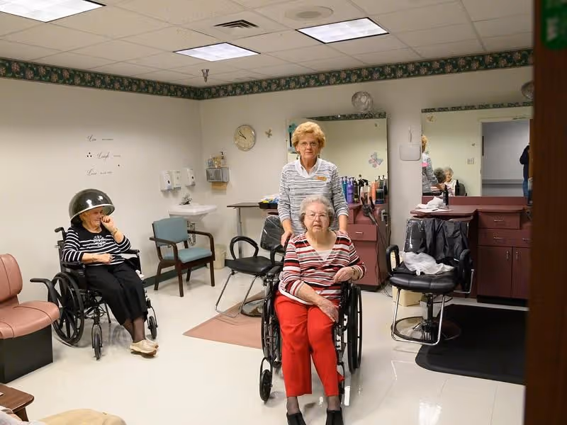 A senior living facility salon room with two elderly women in wheelchairs. One woman is sitting under a hair dryer, and another woman in red pants is being pushed in a wheelchair by a caregiver. The room has salon chairs, mirrors, and hair care products on counters.
