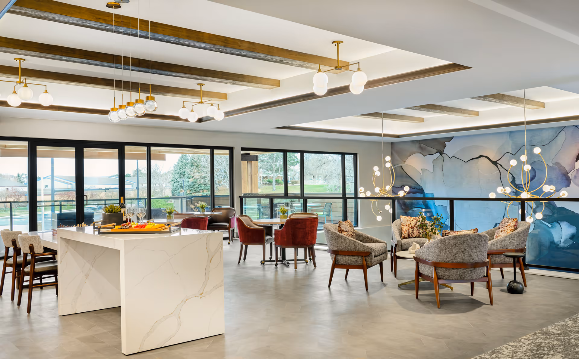 A modern senior living common area with a white marble island table surrounded by chairs, several seating areas with upholstered chairs, large windows providing natural light, and artistic wall decor featuring abstract blue and gray patterns. The ceiling has wooden beams and multiple contemporary light fixtures.