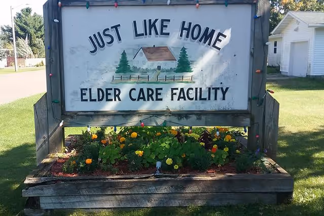 Outdoor sign reading 'Just Like Home Elder Care Facility' featuring a painted house above a flower bed.