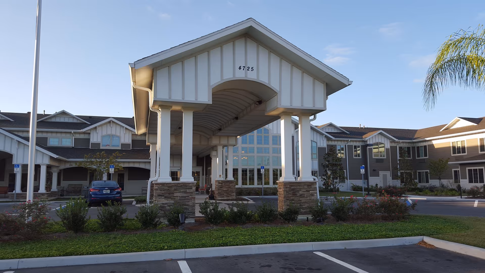 Covered porte-cochère and front facade of a two-story senior living building with landscaping and a parked car.