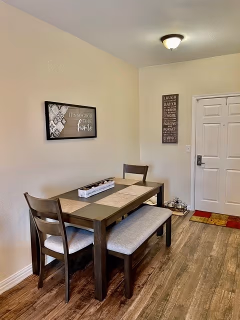 A small dining area with a dark wooden table, two chairs, and a bench with light-colored cushions. The table has placemats and a decorative centerpiece. On the wall above the table is a framed sign that reads 'It's so good to be home.' Next to the table is a white door with a lock and handle, and a vertical wall decoration with inspirational words. The floor is wooden, and the walls are painted light beige.