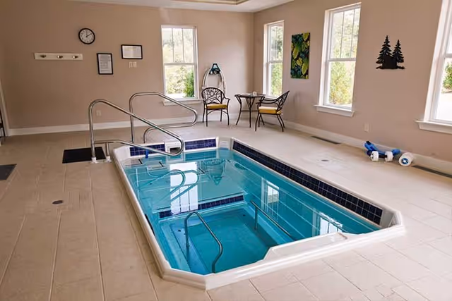 Indoor therapy pool with handrails and steps, surrounded by beige tiled floor. Two chairs and a small round table are placed near the windows, along with some wall decorations and exercise equipment on the floor.