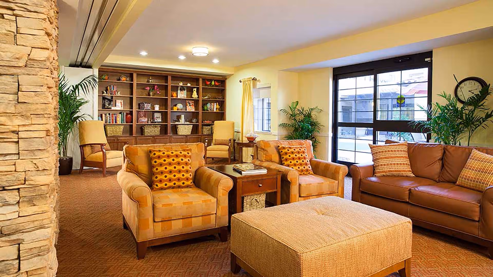 A cozy living room area in a senior living facility featuring two patterned armchairs with orange and yellow cushions, a brown leather sofa with patterned pillows, a large cushioned ottoman, a wooden side table with books, potted plants, a stone accent wall, a large window with black frames, and a wooden bookshelf filled with books and decorative items.
