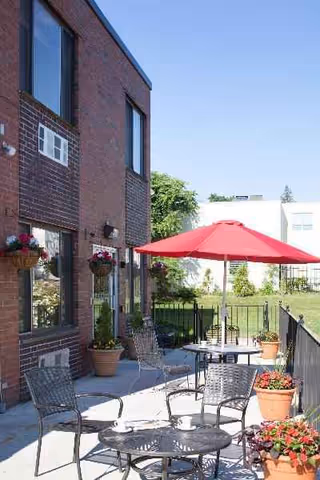 Brick nursing facility exterior with an outdoor patio, seating, planters, and a red umbrella.