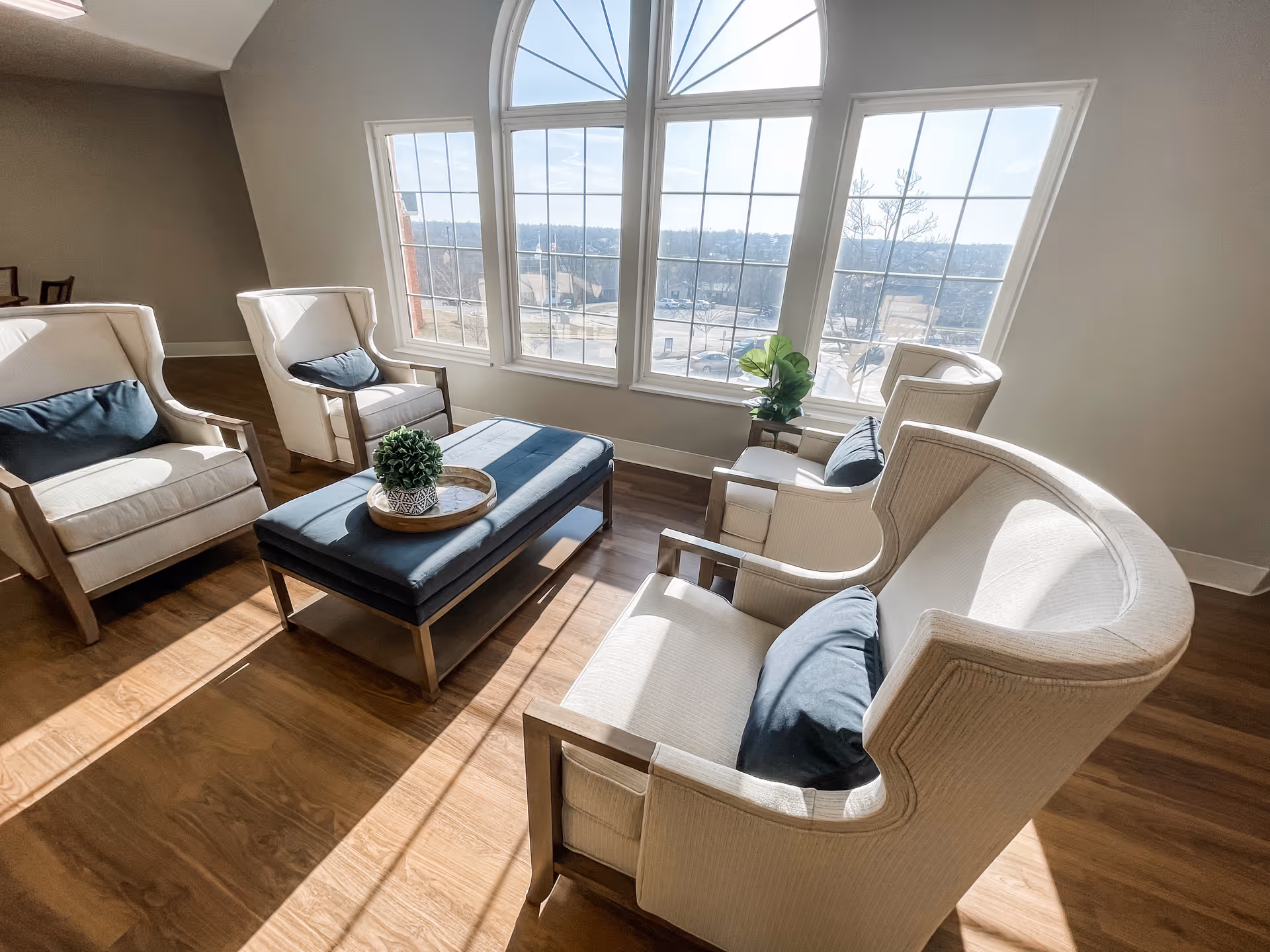 Sunlit sitting area with upholstered armchairs and a central upholstered ottoman in front of large arched windows.