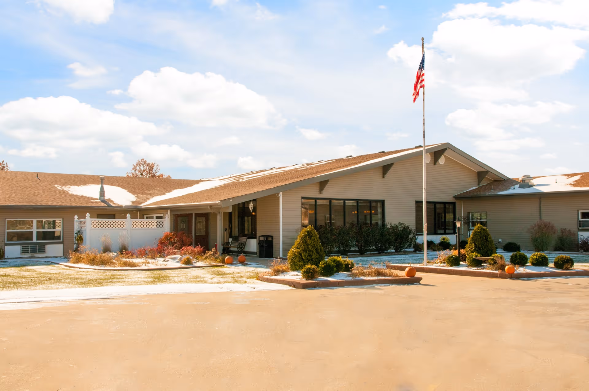 Front exterior of a single-story senior living facility with a flagpole, landscaping, and pumpkins by the entrance.