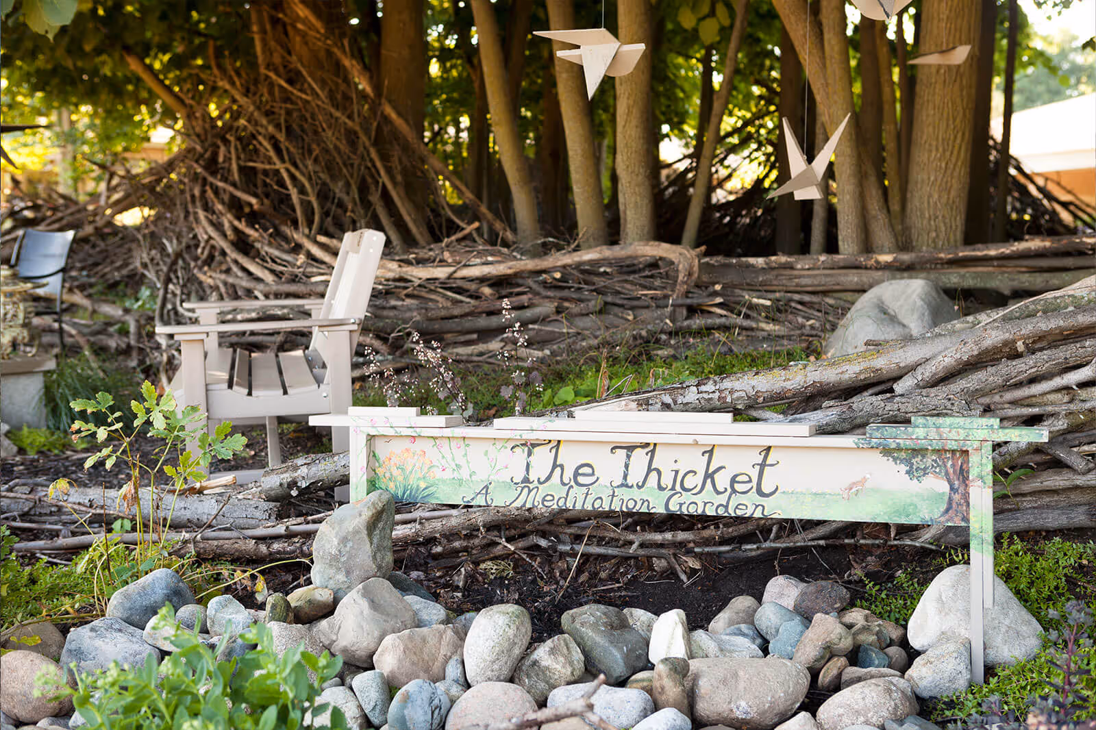 Shaded outdoor meditation garden with Adirondack chairs, a painted wooden sign reading "The Thicket — A Meditation Garden," rocks and trees.