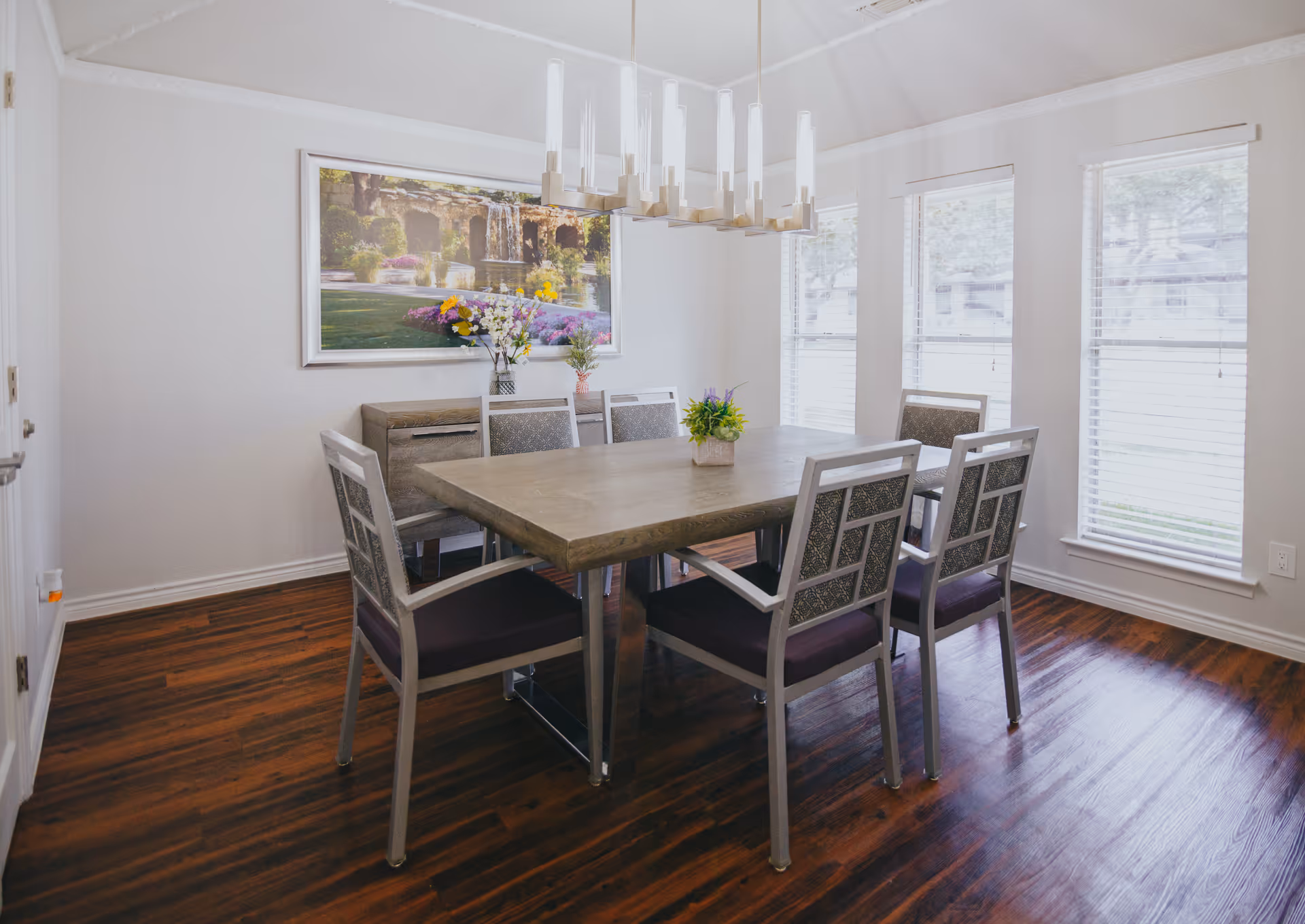 Bright dining room with a rectangular wooden table surrounded by six chairs, a modern chandelier overhead, and large windows letting in natural light.