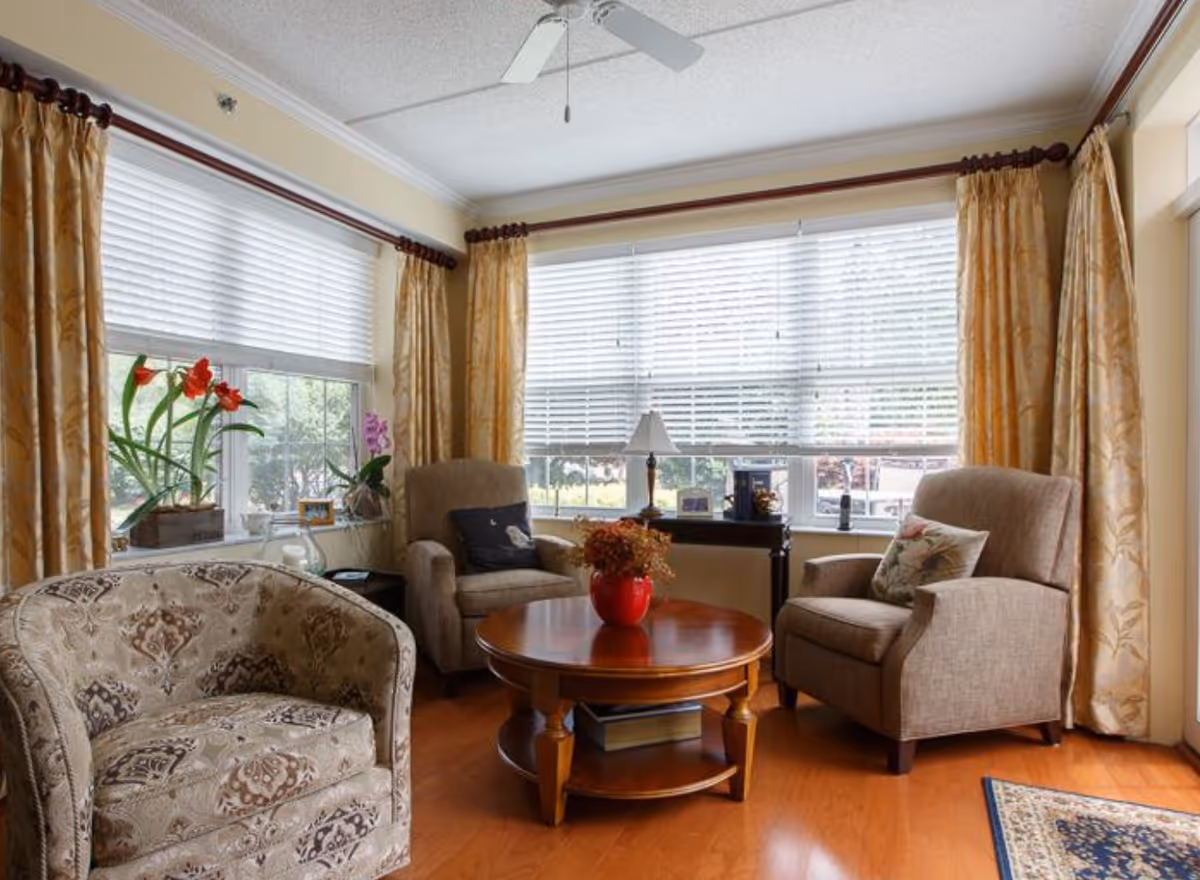 Sunny living room with upholstered chairs around a round wooden coffee table, large windows with blinds and gold curtains, and potted plants on the windowsill.