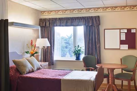 Bedroom in a senior facility with a burgundy bedspread, window, plants, and a table with chairs.