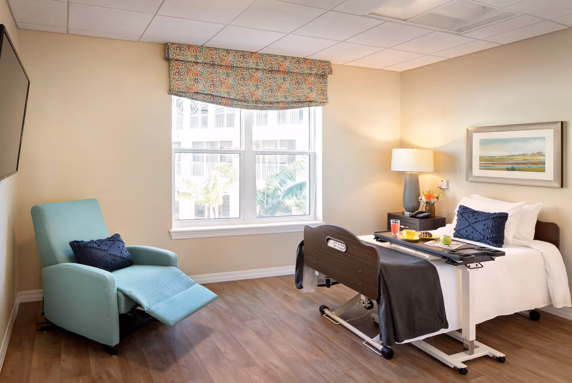 Bright senior living bedroom with a hospital-style bed and overbed tray, a teal recliner, window, lamp, and framed artwork.