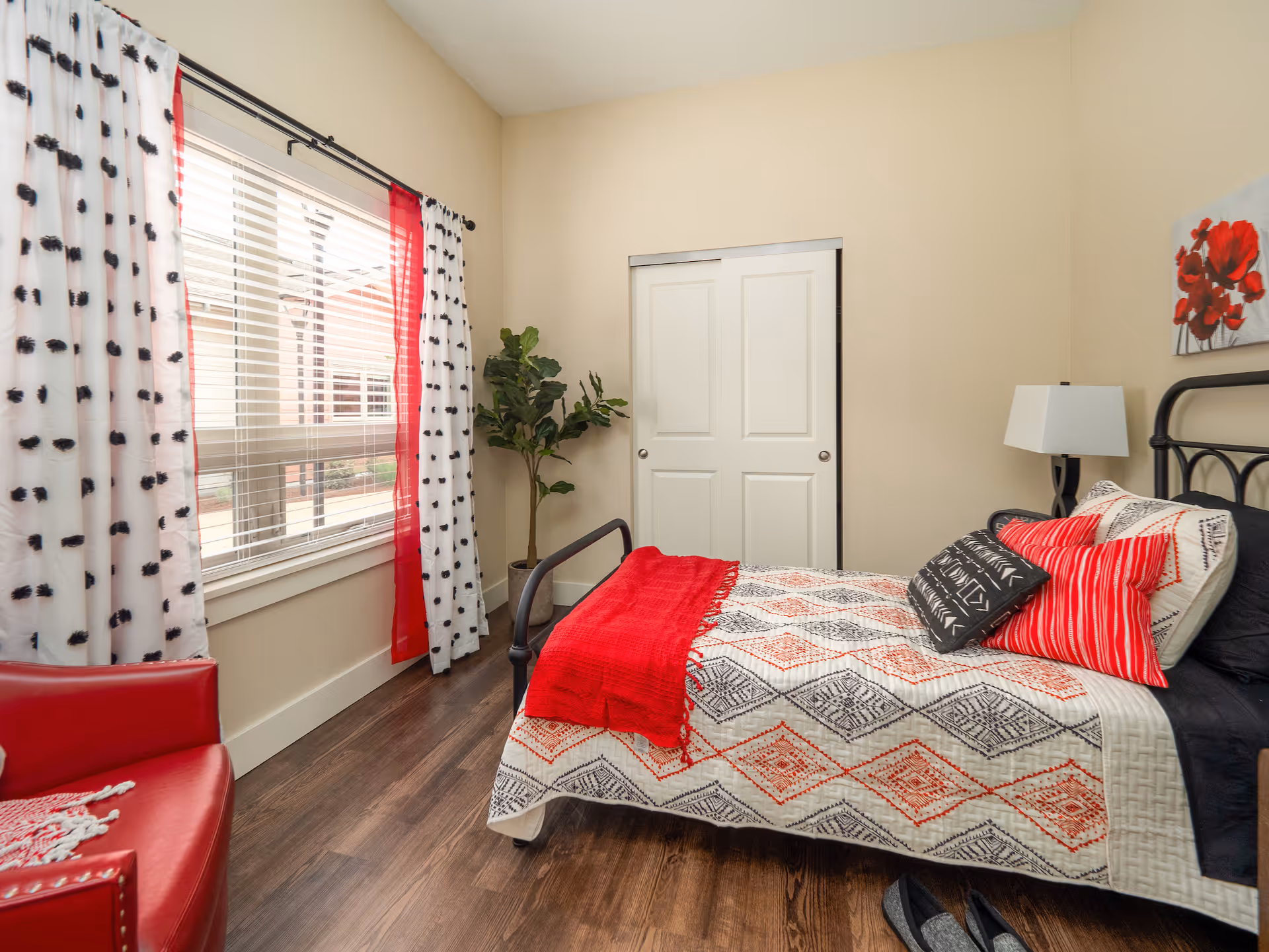 A cozy bedroom with a single bed featuring a patterned quilt and red throw blanket, accented with red and black pillows. There is a white lamp on a bedside table, a red armchair, a potted plant near a window with white and red curtains, and a closet with sliding white doors.