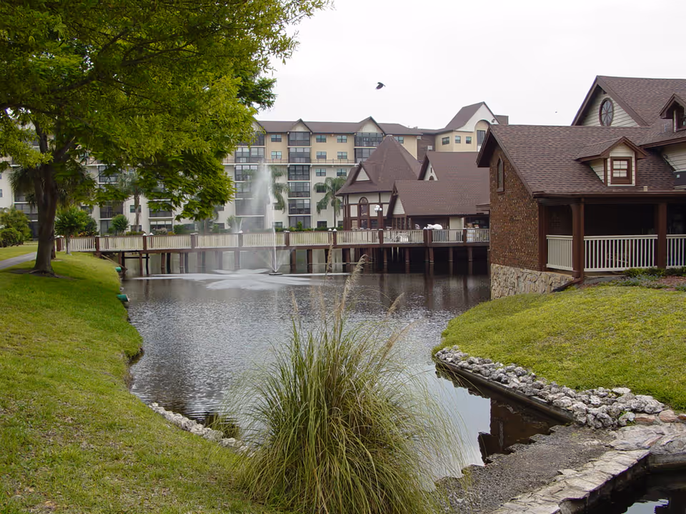 A serene outdoor scene at Freedom Village of Bradenton featuring a pond with a water fountain in the center. Surrounding the pond are green grassy areas, trees, and a wooden bridge connecting buildings with brown roofs and white railings. In the background, there is a multi-story residential building with balconies.