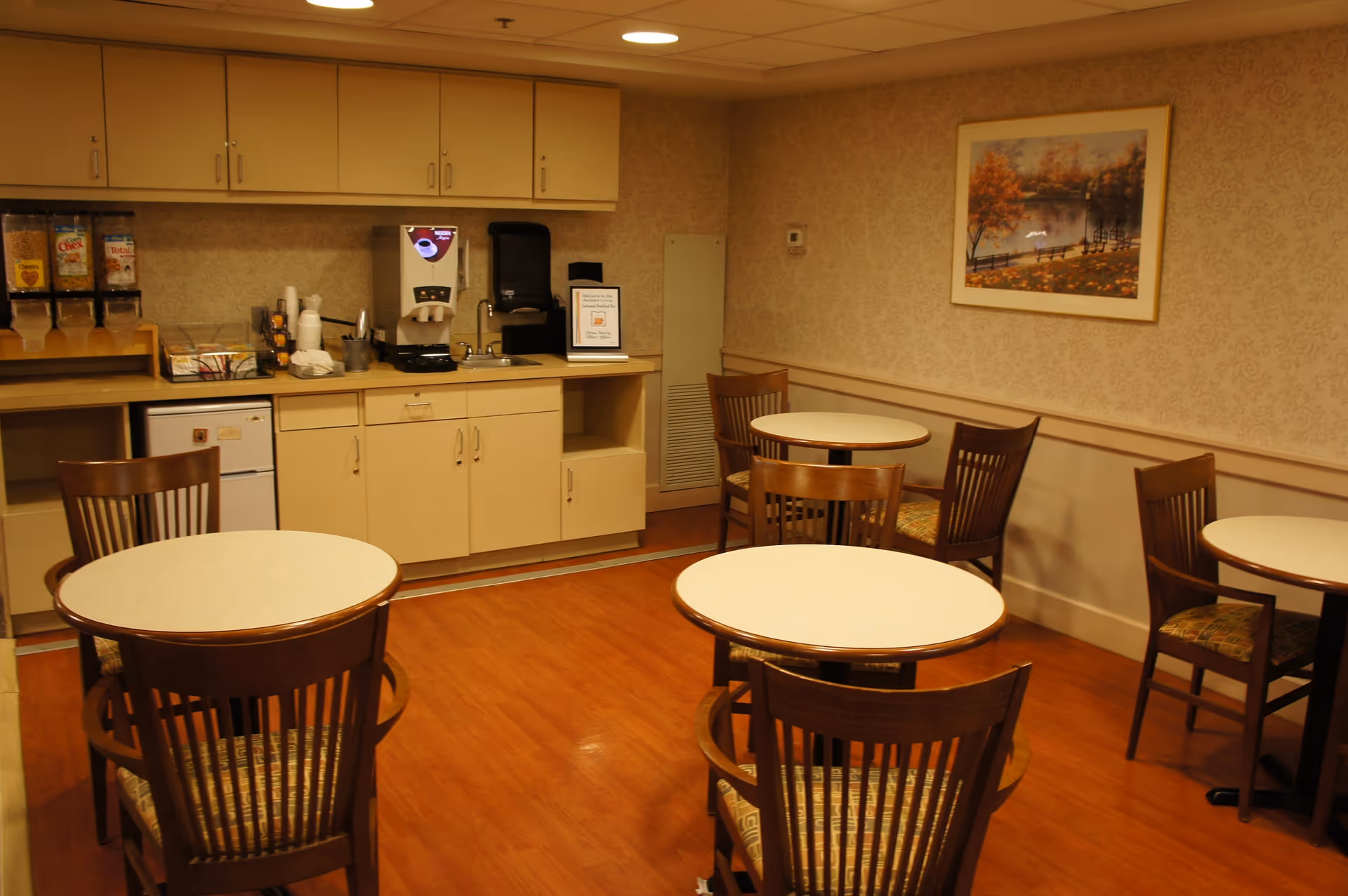 Small dining area with round tables and wooden chairs and a kitchenette with a coffee dispenser along the back wall.