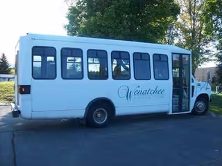 A white shuttle bus parked in Kadie Glen's lot beside trees.