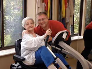 An elderly woman sitting on a recumbent exercise bike inside a room, smiling and holding the bike handles, with a man in a red shirt standing behind her, also smiling. Large windows with a view of greenery are visible in the background.