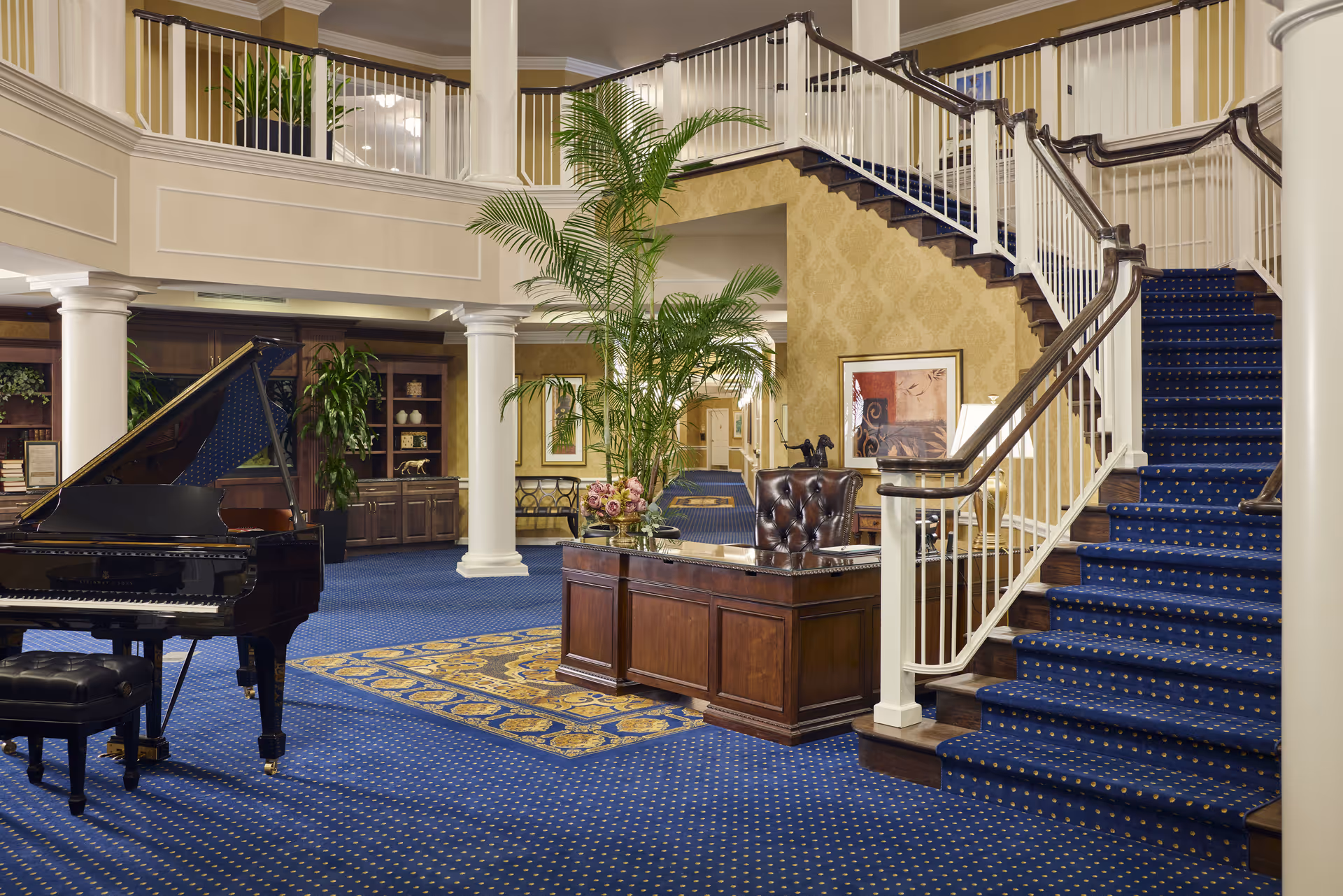 Elegant interior of a senior living facility featuring a grand piano on a blue carpet with gold patterns, a wooden reception desk with a leather chair and floral arrangement, white columns, a staircase with blue carpet and white railing, and decorative plants and framed artwork on the walls.