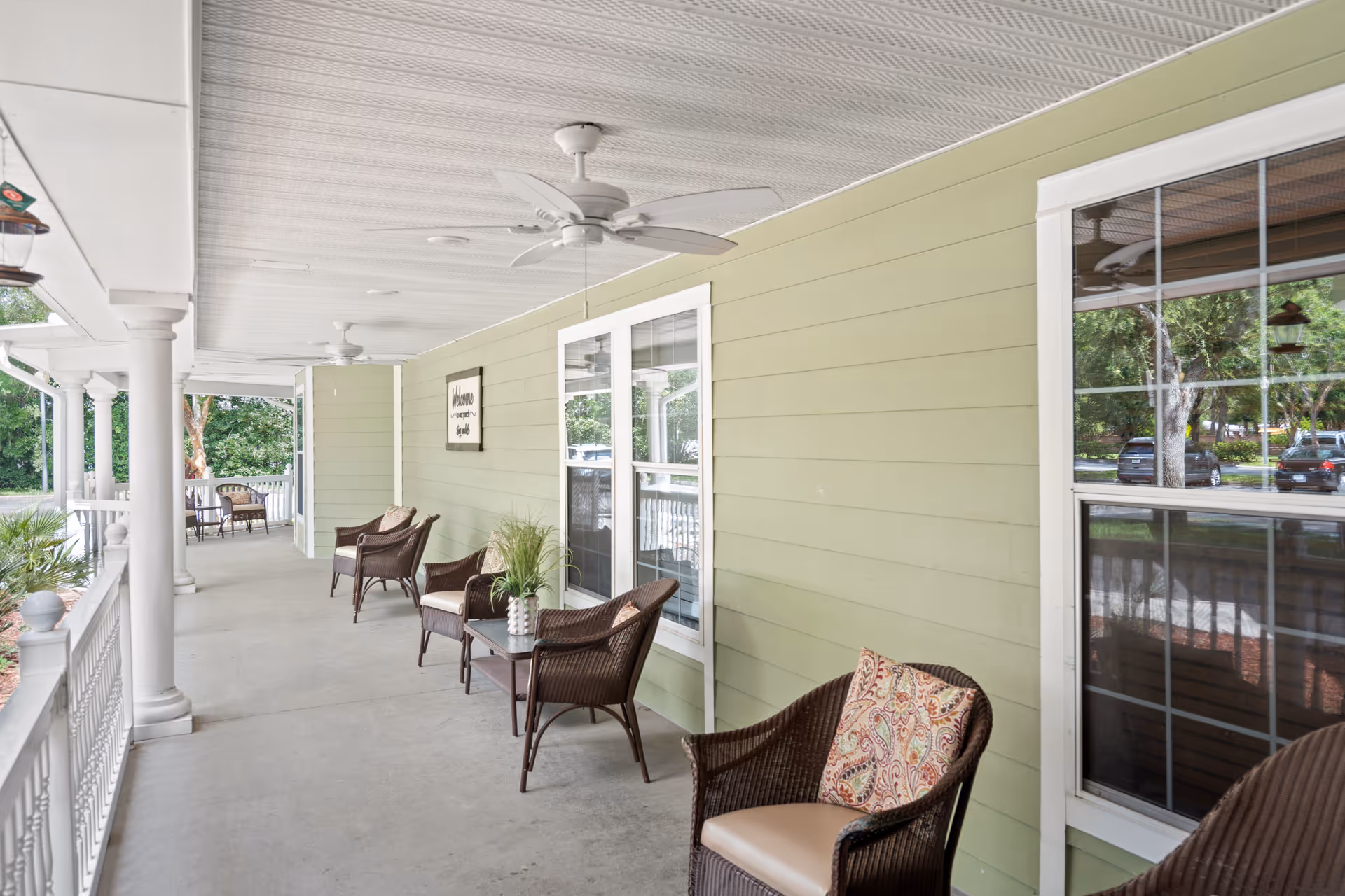 A spacious covered porch with light green siding and white trim, furnished with wicker chairs and small tables. Ceiling fans are installed overhead, and the porch overlooks a landscaped area with trees and parked cars visible in the background.