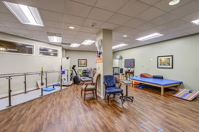 A spacious therapy and exercise room with parallel bars, fitness equipment, a treatment table, chairs, and wood flooring.