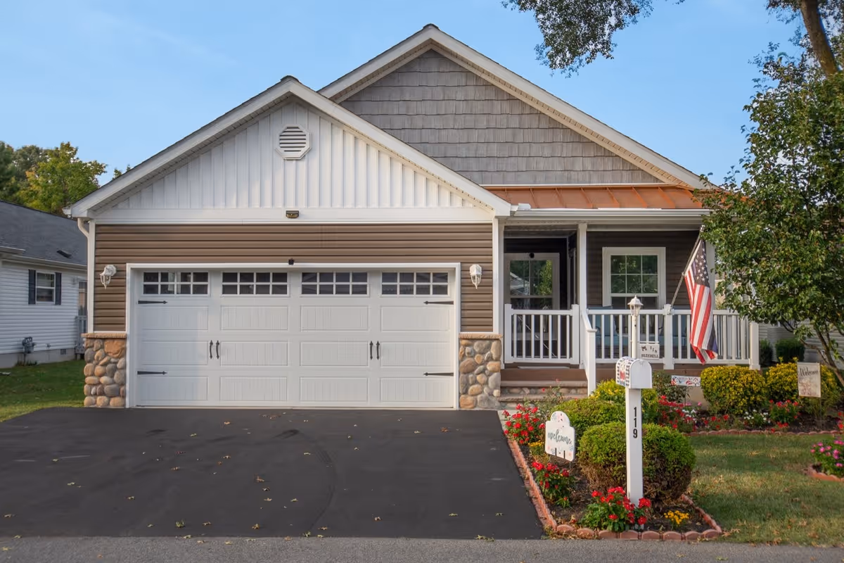 Front exterior view of a single-story house with a two-car garage, a small porch with white railing, an American flag, a mailbox with the number 119, and a well-maintained garden with flowers and shrubs.