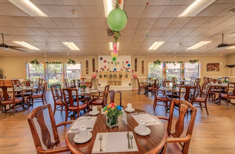 A spacious dining room in a senior living facility with multiple wooden tables and chairs arranged neatly. Each table is set with placemats, cups, and utensils. The room is decorated with colorful hanging paper lanterns and floral wall decorations. Large windows allow natural light to brighten the space, and there are plants and flowers on the tables and windowsills.