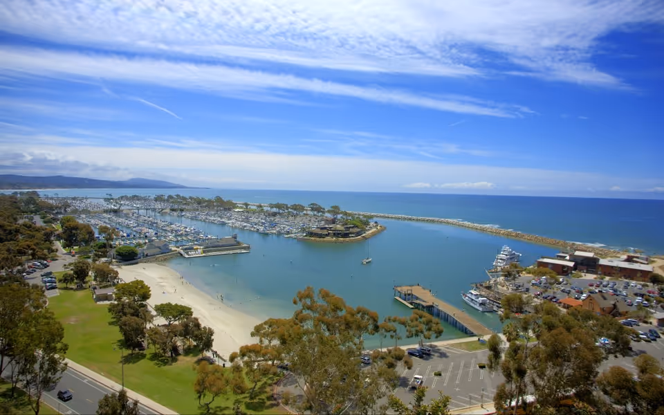 Aerial view of a coastal marina with numerous boats docked in the water, a sandy beach area, green parkland with trees, parking lots, and buildings under a partly cloudy blue sky.