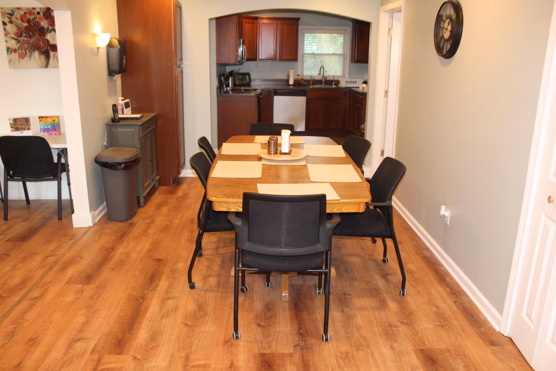 Dining area with a wooden table set with placemats and black chairs, opening into a kitchen.