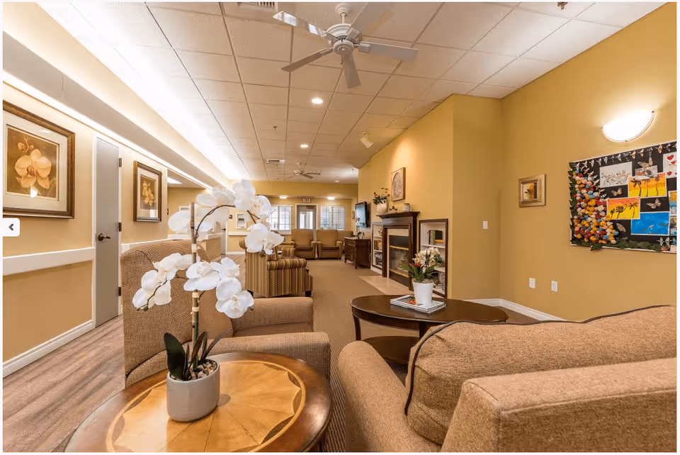 A warm, well-lit senior living common room with upholstered chairs, coffee tables, a fireplace, and a bulletin board.