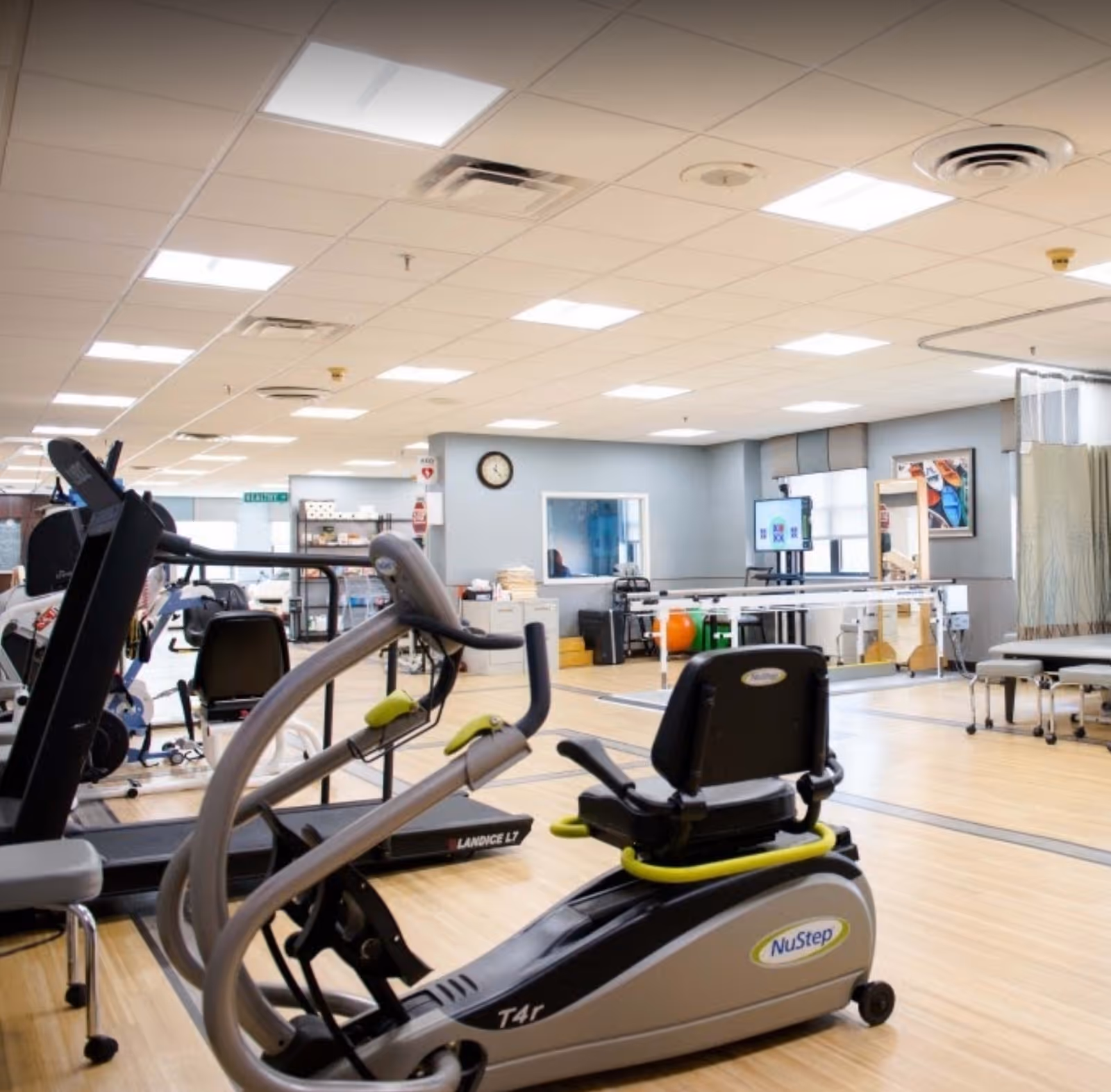 A spacious rehabilitation and exercise room with various fitness equipment including a NuStep recumbent cross trainer and treadmills. The room has light wood flooring, a drop ceiling with fluorescent lights, and light blue walls. There are therapy tables, a clock on the wall, and a TV screen mounted in the background.